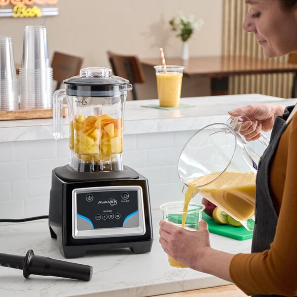 A commercial blender with a digital keypad control and a clear container filled with chopped fruit, shown on a countertop with a person pouring a blended drink into a cup.