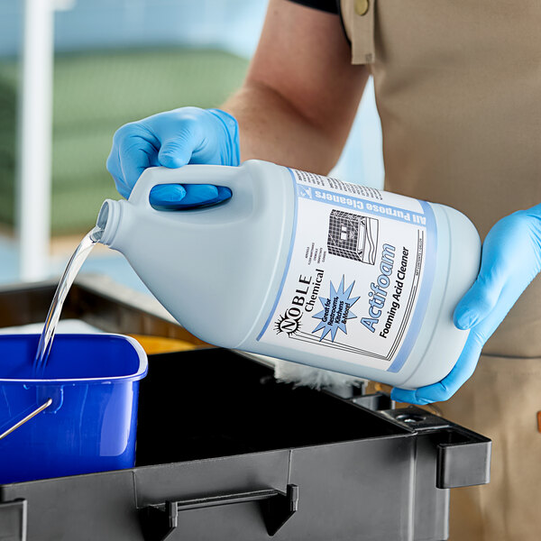 A person in blue gloves pouring Noble Chemical Actifoam concentrated restroom cleaner into a white container.