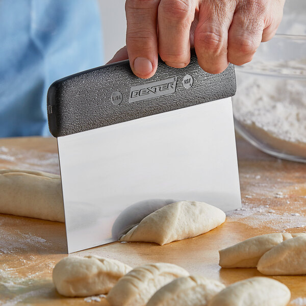 A person using a Dexter-Russell bench scraper to cut dough on a table.