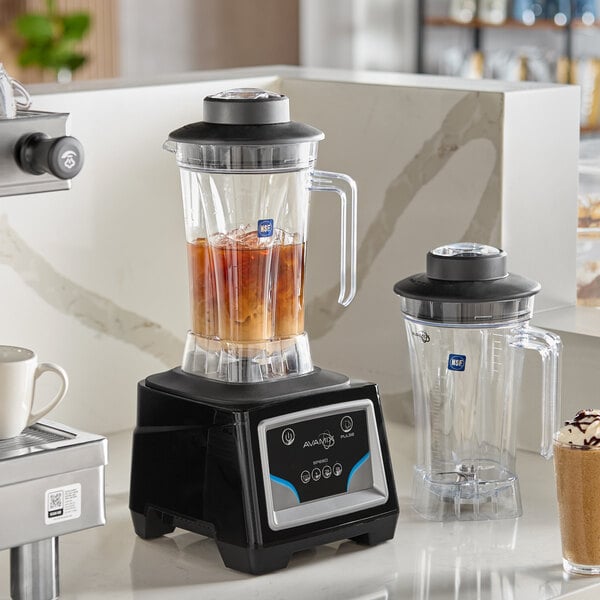 A commercial blender with a keypad control base and two large clear containers, one filled with iced liquid and the other empty, both with handles and lids.