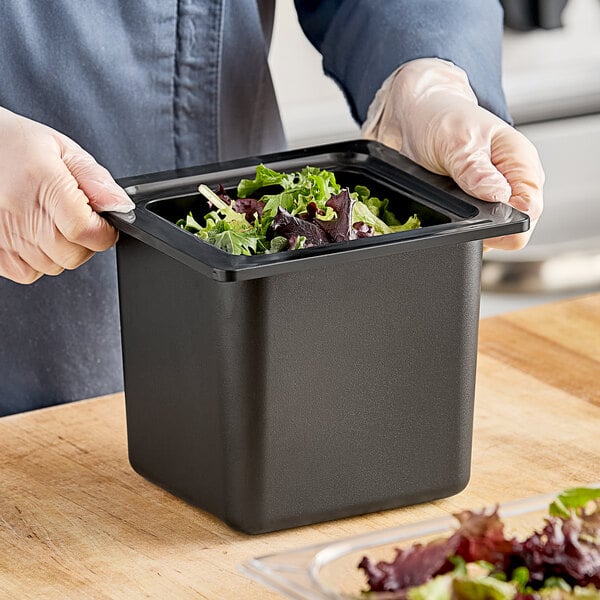 A person in gloves holding a San Jamar black food pan filled with food on a counter.
