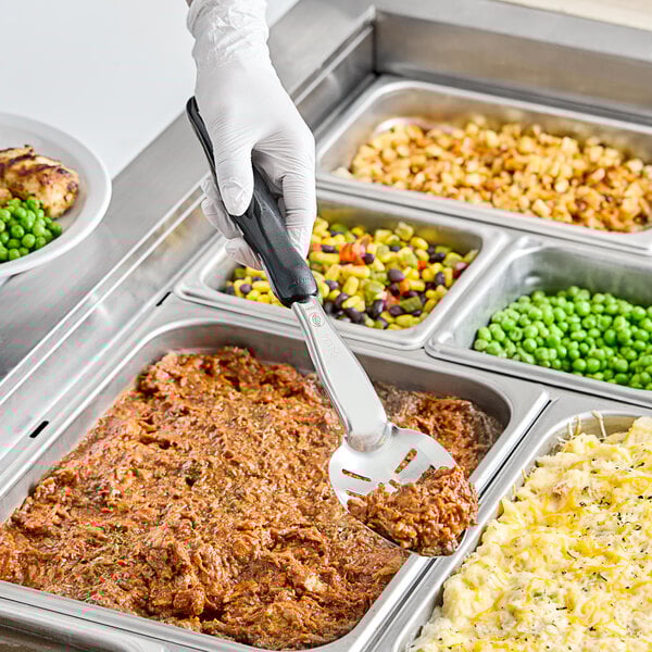 A hand holding a Vollrath Jacob's Pride slotted basting spoon with a black handle over food.