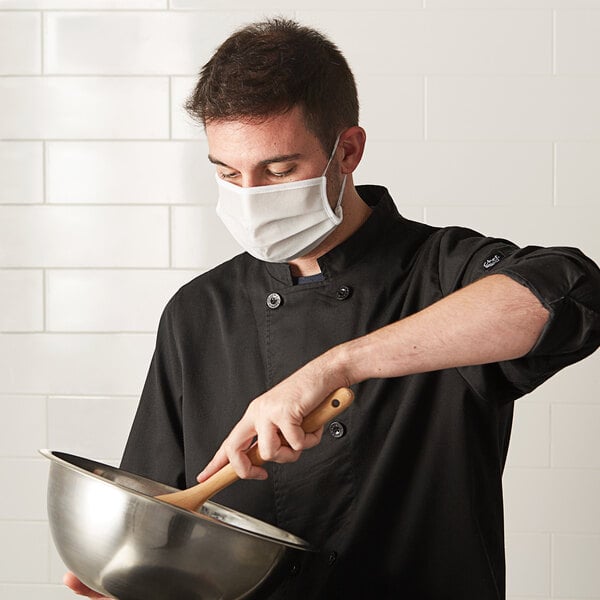 A man wearing a Mercer Culinary white protective face mask while mixing a bowl with a wooden spoon.