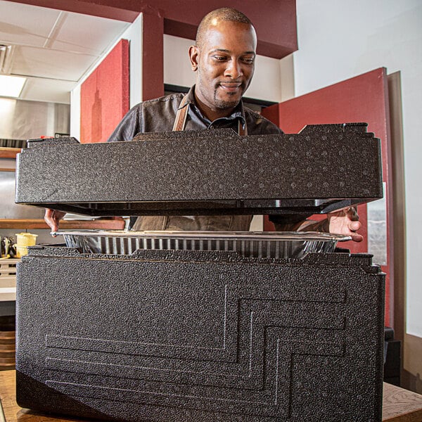 A man standing in a kitchen holding a large black Cambro container.