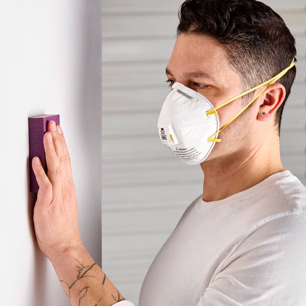 A person wearing a 3M 8210V N95 particulate respirator with a Cool Flow valve while sanding a wall.