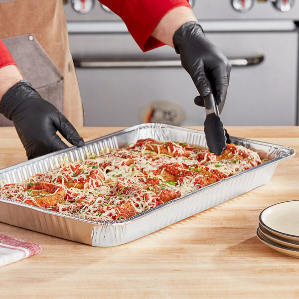 A person in black gloves cutting pizza in a Choice foil steam table pan on a table.