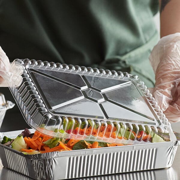 A person in gloves holding a Choice oblong foil take-out container of food.