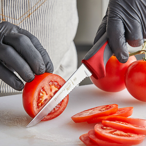 A person in a black glove using a Dexter-Russell scalloped utility knife to cut a tomato.