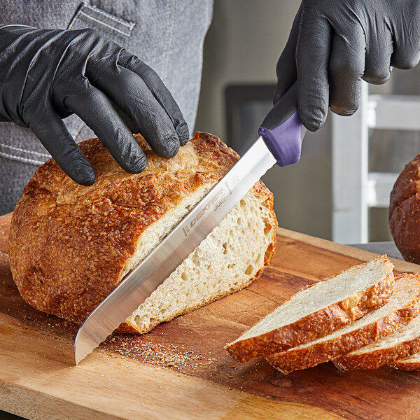 A person in black gloves using a Dexter-Russell scalloped bread knife to cut a loaf of bread.