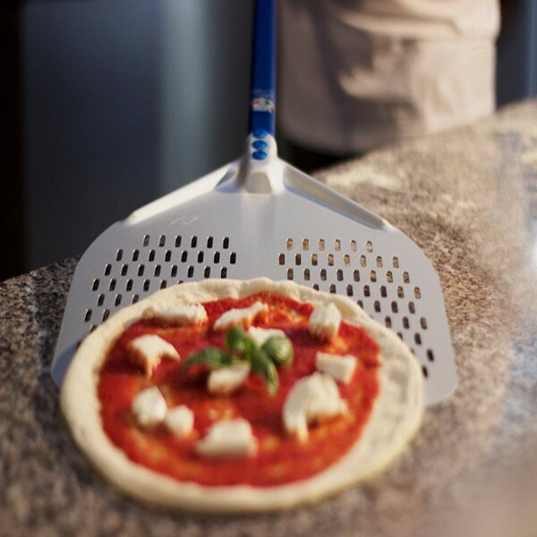 A pizza with tomato sauce, cheese, and basil is placed on a perforated metal pizza peel with a blue handle, resting on a countertop.