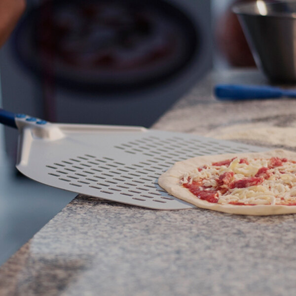A metal pizza peel with a perforated surface is being used to lift an uncooked pizza from a countertop.