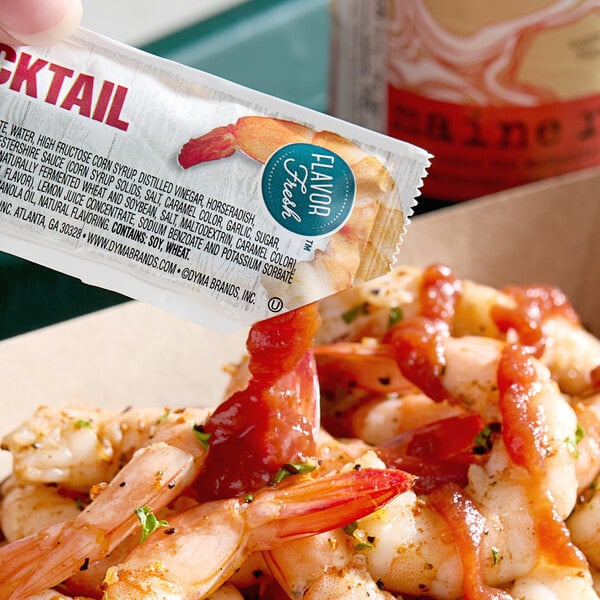 A hand holding a Cocktail Sauce packet over shrimp.