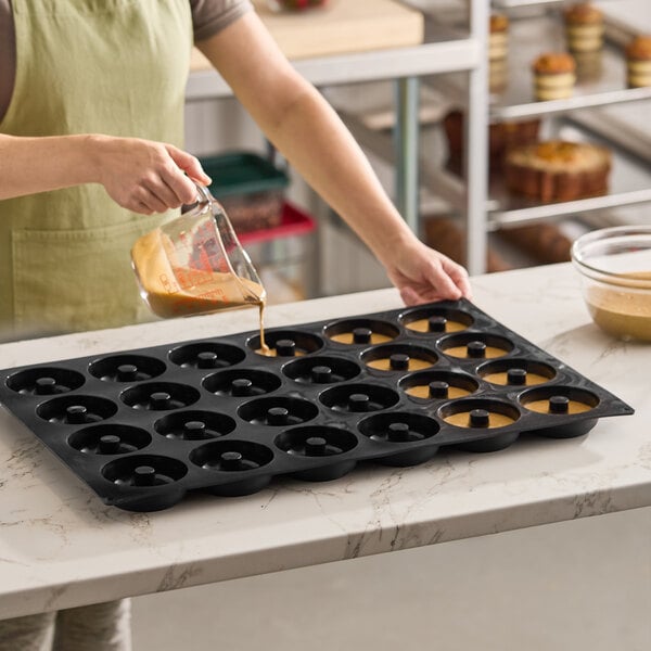A black silicone baking mold with 24 donut-shaped compartments being filled with batter.