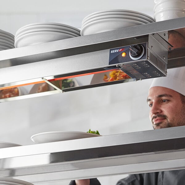 A chef in a professional kitchen adjusts the temperature on a ServIt strip warmer above a shelf with white plates.