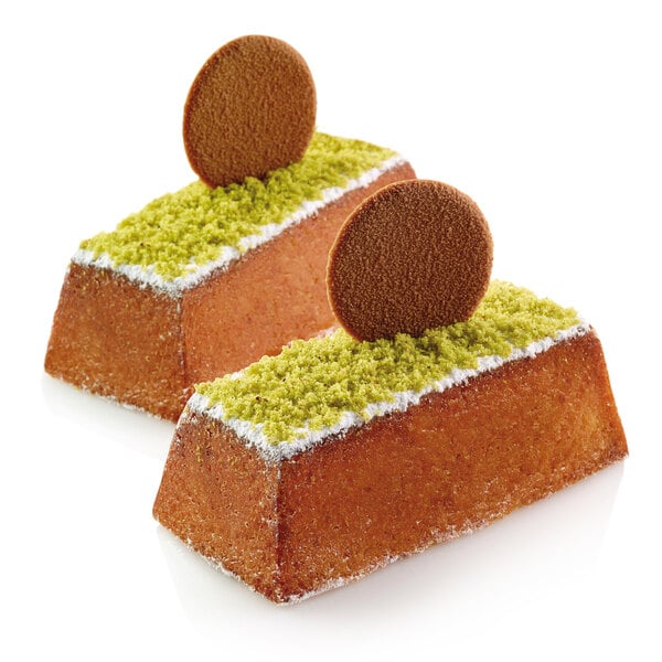 Two Silikomart mini cakes with green icing and a cookie on top displayed in a bakery.