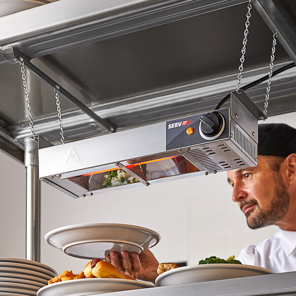 A man holding a plate of food under a ServIt strip warmer on a table in a professional kitchen.