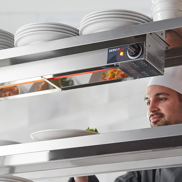 A chef in a professional kitchen looks at a stack of white plates on a shelf above a ServIt strip warmer.