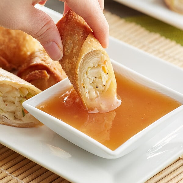 A hand dipping a fried egg roll into a bowl of Lee Kum Kee Gold Label Plum Sauce.