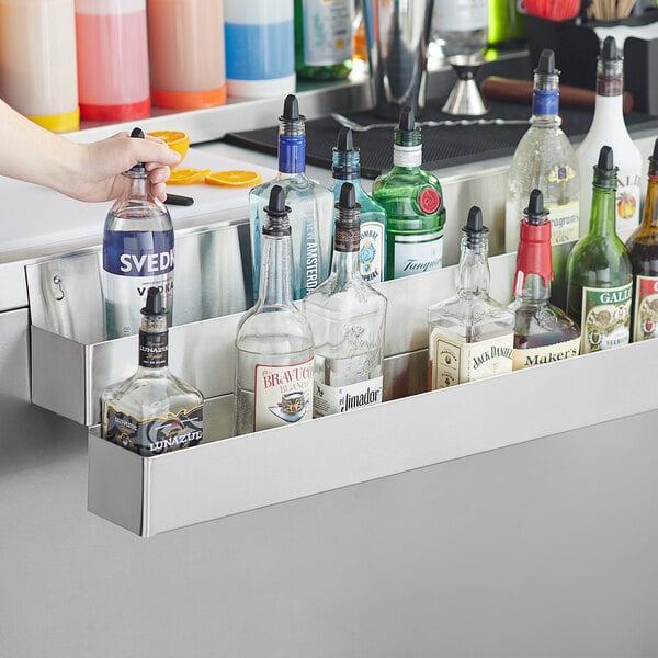 A person using a Regency stainless steel double tier speed rail to hold a bottle of alcohol on a bar counter.