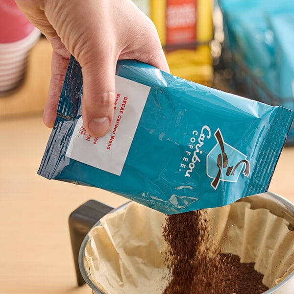 A hand holding a blue Caribou Coffee package pouring brown Caribou Coffee grounds into a coffee maker.