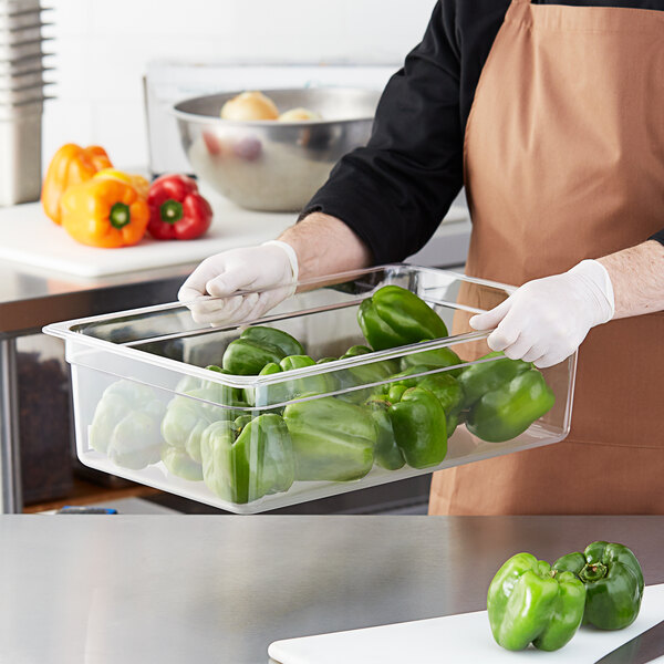 A person in a chef's uniform holding a clear polycarbonate food pan filled with yellow, red, and green peppers.