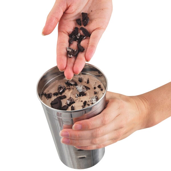 A person pouring cookies into a cup of milkshake using a Hamilton Beach drink mixer.