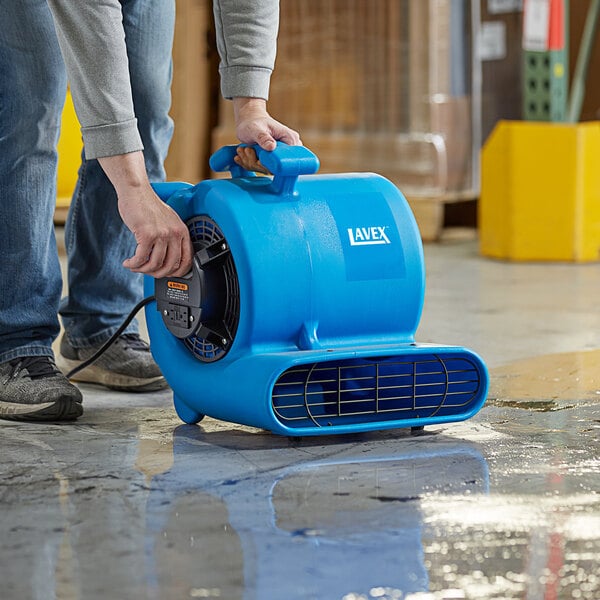 A person using a Lavex blue air blower to clean a floor in a room.