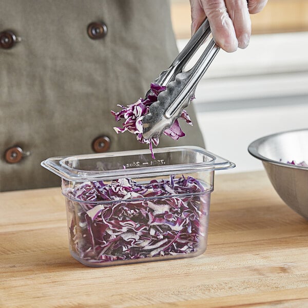 A person using tongs to add shredded cabbage from a clear plastic food pan to a bowl.