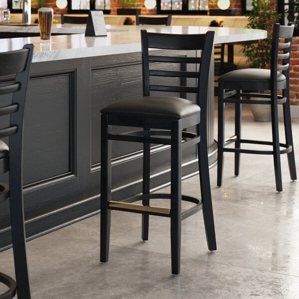 A Lancaster Table & Seating black wood bar stool with black vinyl seat at a table.