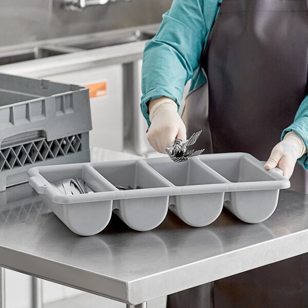 A woman in a blue apron holding a white Rubbermaid container with four sections of silverware in it.