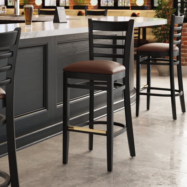 A Lancaster Table & Seating black wood ladder back bar stool with a dark brown vinyl seat on a black surface.