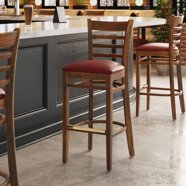 A Lancaster Table & Seating wooden ladder back bar stool with a burgundy vinyl seat next to a table in a restaurant.
