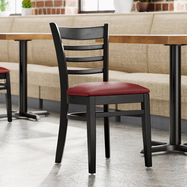 A Lancaster Table & Seating black wood ladder back chair with a burgundy vinyl seat next to a table.