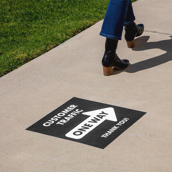 A person walking on a sidewalk next to grass with black boots and a black square floor decal that says "Customer One Way" in white text.