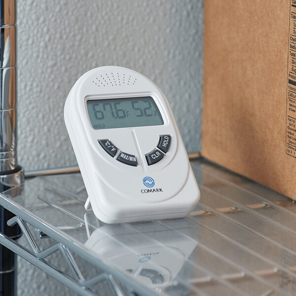 A Comark DTH880 digital indoor thermometer and hygrometer on a glass shelf with a screen.
