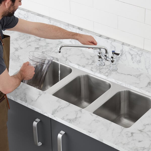 A person pouring water into a stainless steel three compartment sink on a white counter top.