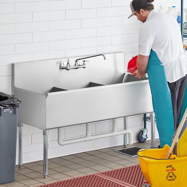 A man in a blue apron cleaning a Regency stainless steel utility sink.