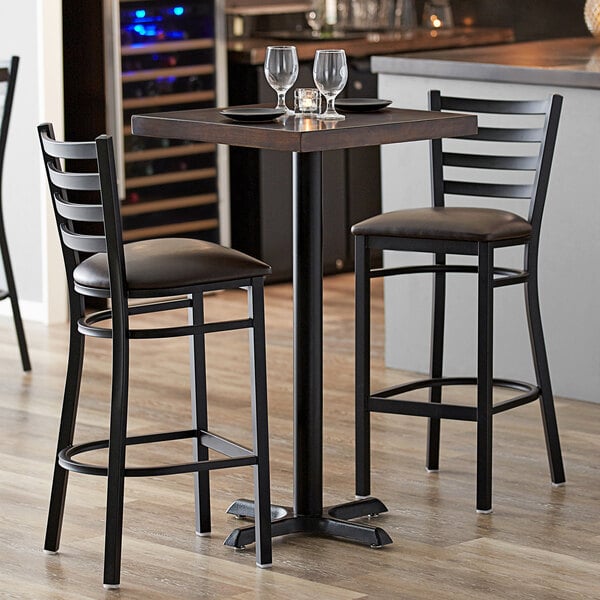 A Lancaster Table & Seating square bar height table with espresso finish and cast iron base plate with two chairs and wine glasses on it.