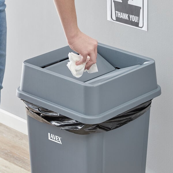 A woman cleaning a Lavex gray square trash can with a paper towel.