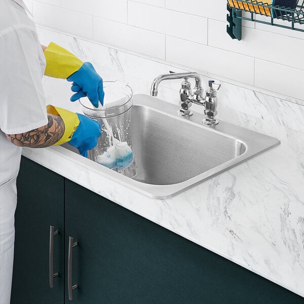 A man in blue gloves washing dishes in a Waterloo stainless steel drop-in sink.