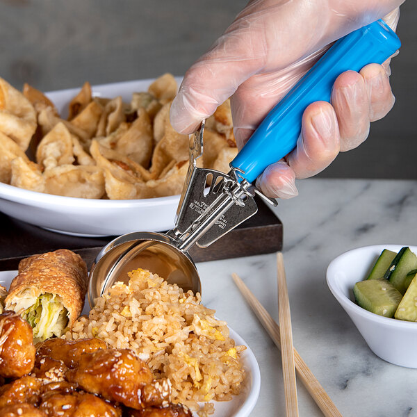 A hand holding a Carlisle blue thumb press disher with a spoon over a bowl of food.