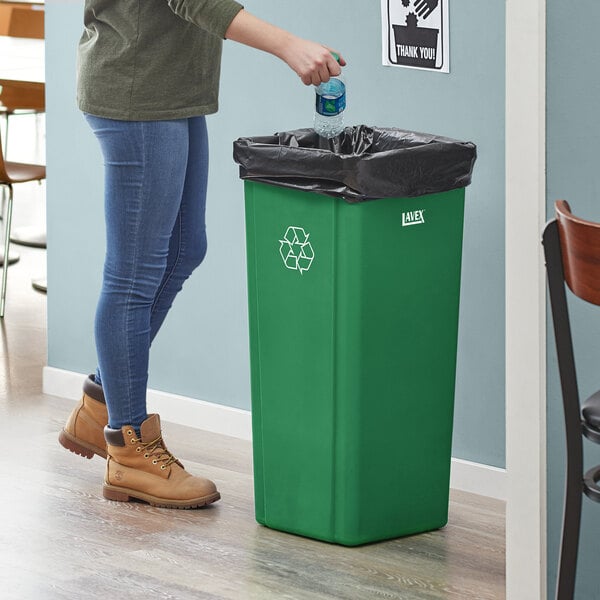 A woman putting a bottle in a Lavex green square recycle bin with a recycle symbol.