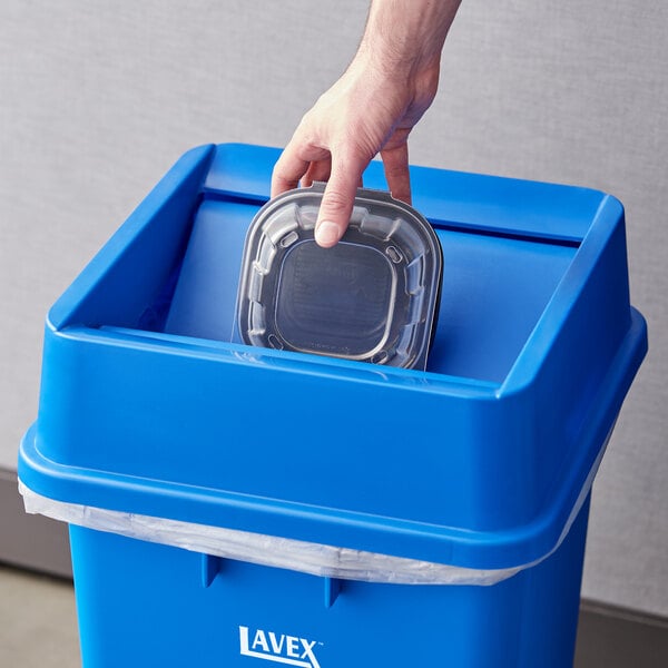 A hand putting a plastic container in a Lavex blue trash can with a swing lid.