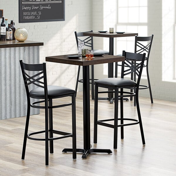 A Lancaster Table & Seating bar height table with two black chairs with black cushions.