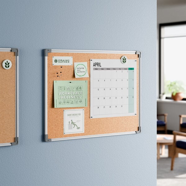 A 24 by 18 inch wall-mount cork board with an aluminum frame, displayed with various notes and a calendar.