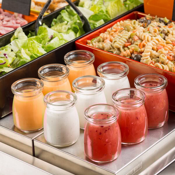A Tablecraft glass jar filled with orange liquid on a table with a group of jars of different colored sauces and pasta salads.