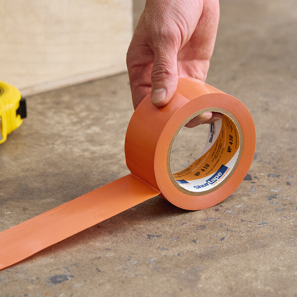 A roll of Shurtape VP 410 orange line set tape being applied to a concrete surface.