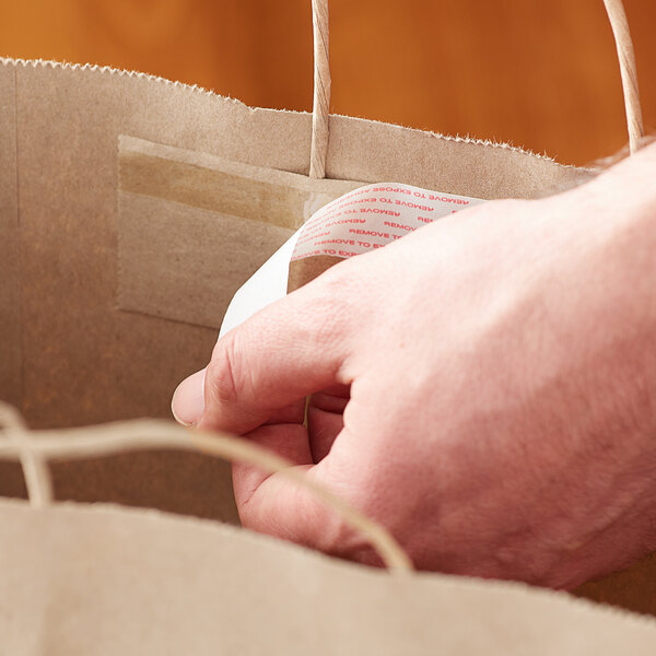 A hand holding a Sabert 2 meal kraft paper bag with a label taped to it.