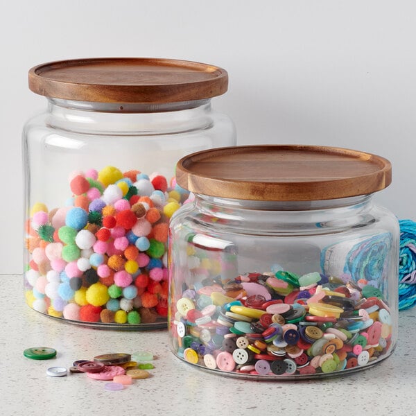 A clear glass jar with a wooden acacia lid, shown filled with colorful craft supplies.