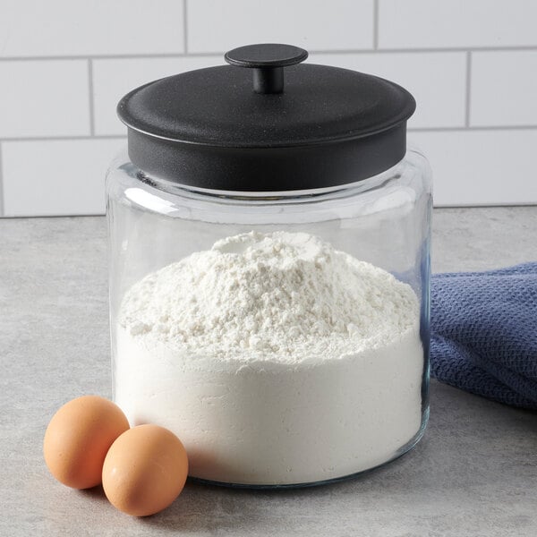 A clear glass jar with a black lid filled with flour, placed next to two eggs and a blue cloth.